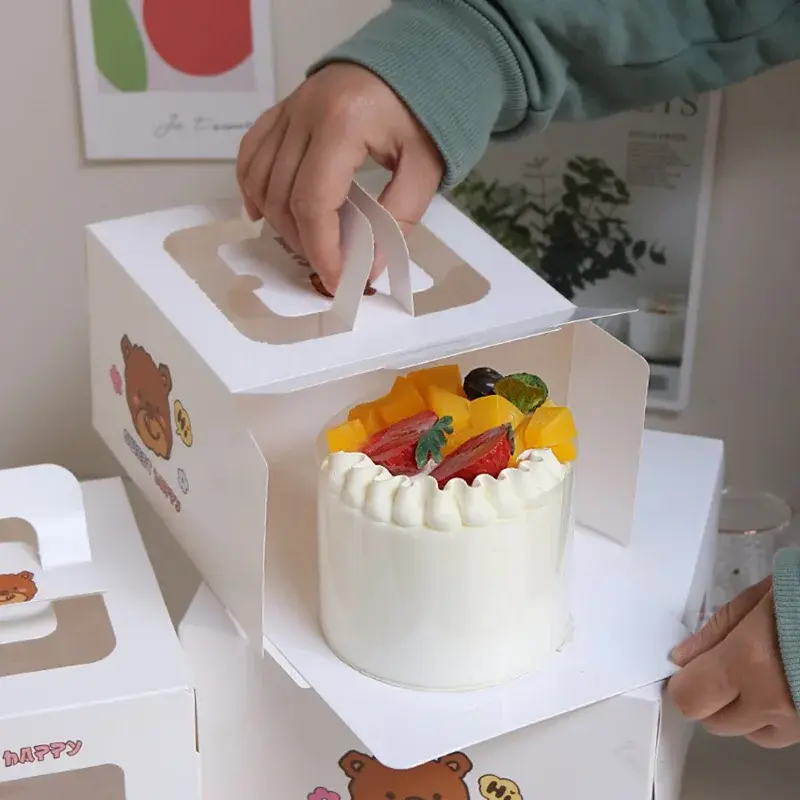 Hand lifting the handle of a white window box containing a cake.